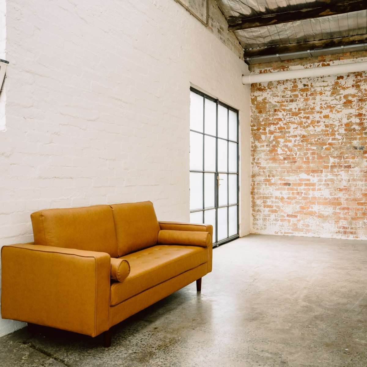 A rust-colored sofa sits against a brick wall in a spacious, industrial-style interior with large windows.