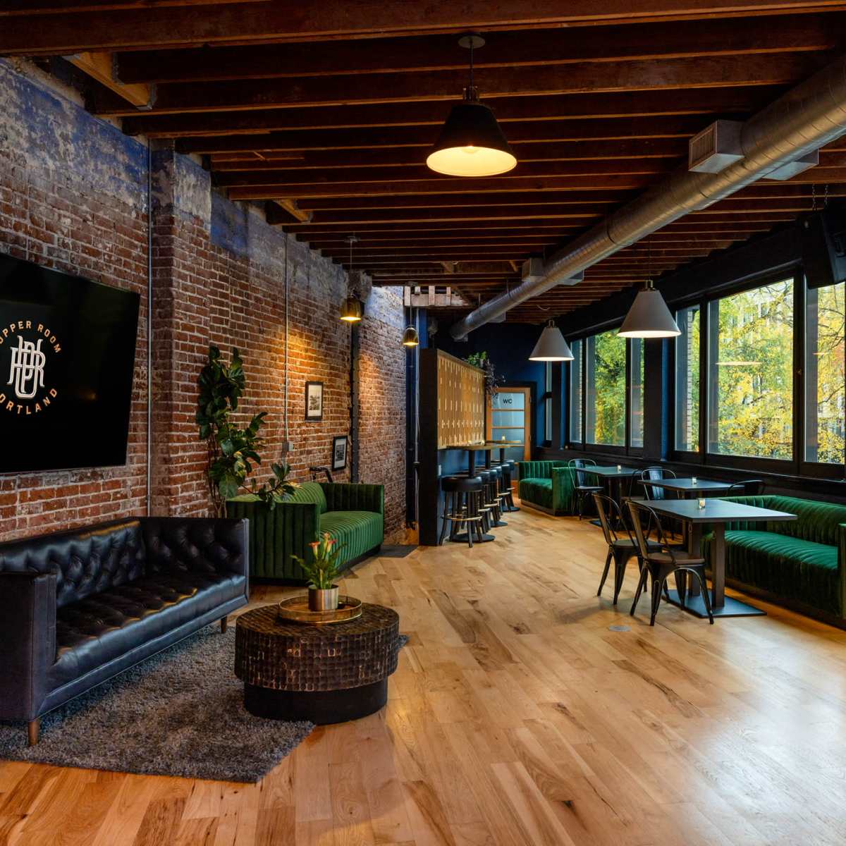 The interior features exposed brick walls, wooden beams, and a mix of seating areas, with large windows providing natural light and a view of trees.