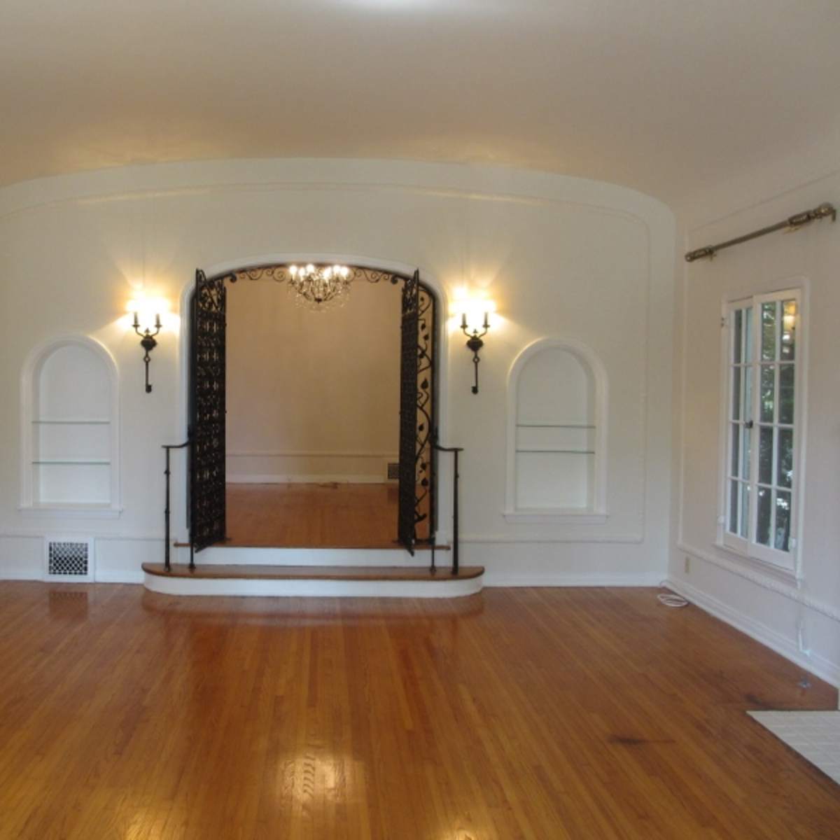 A spacious room featuring an archway with decorative ironwork, two wall sconces, and hardwood flooring.