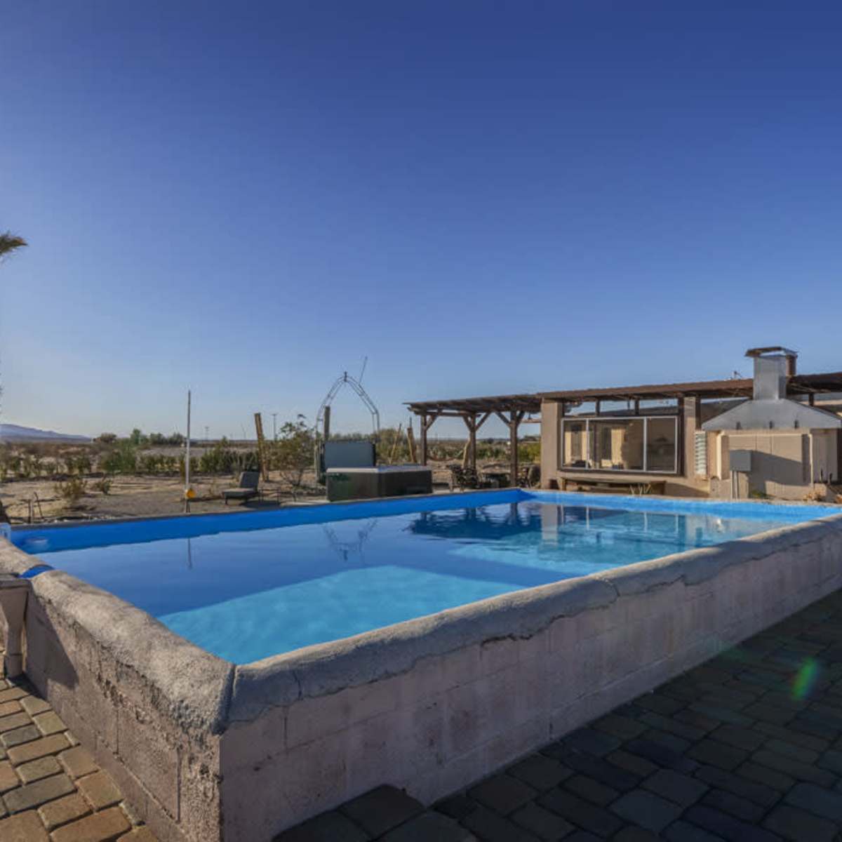 A swimming pool surrounded by patio stones and a palm tree, with solar panels and a clear blue sky in the background.