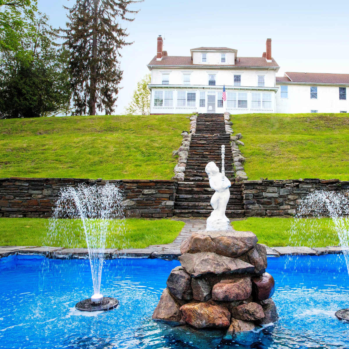 A large white house on a hill, with a stone fountain and blue water in the foreground, and a pathway leading up to the house.