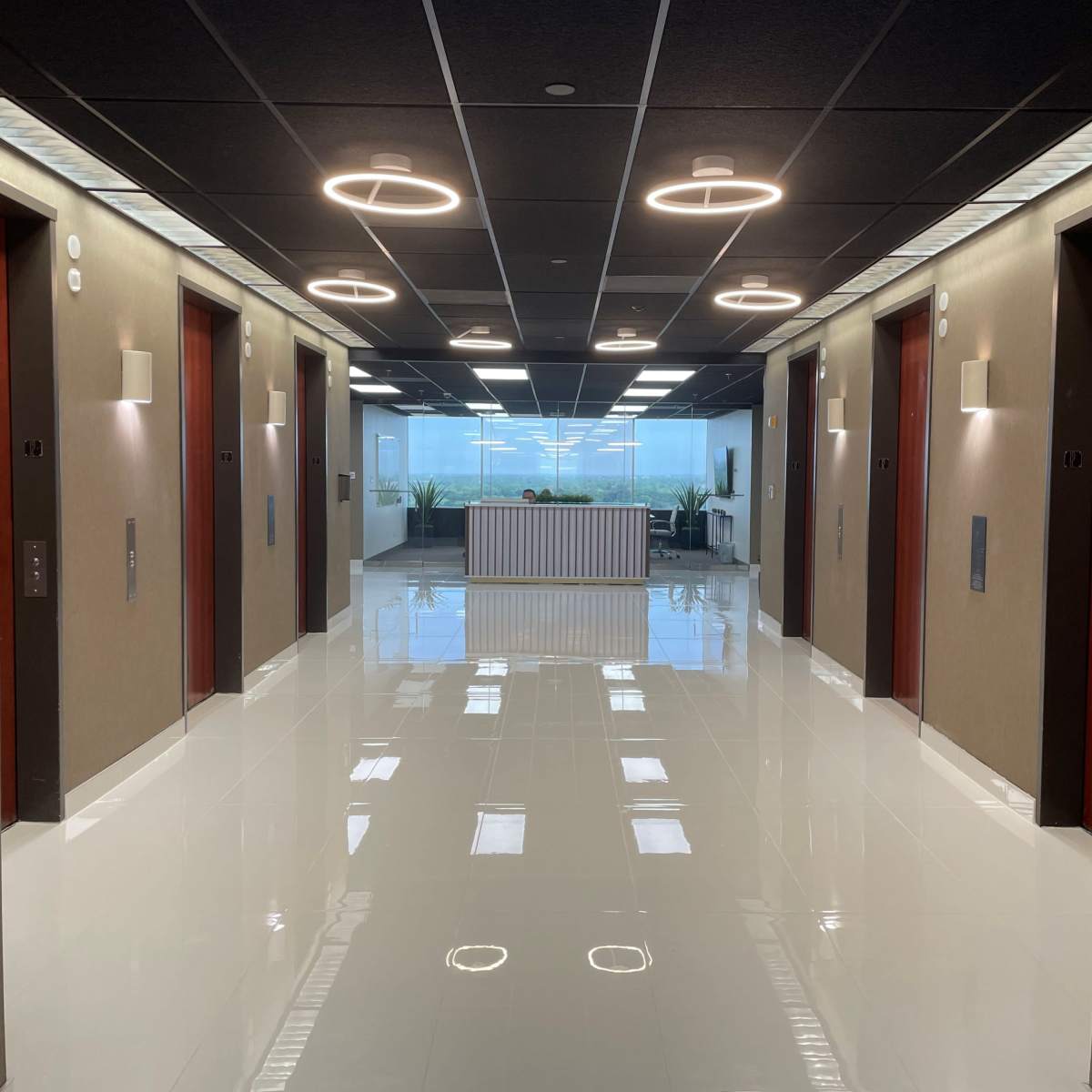 A hallway features multiple glass-walled elevator doors and bright overhead lighting, leading to a lobby area with plants and a reception desk.
