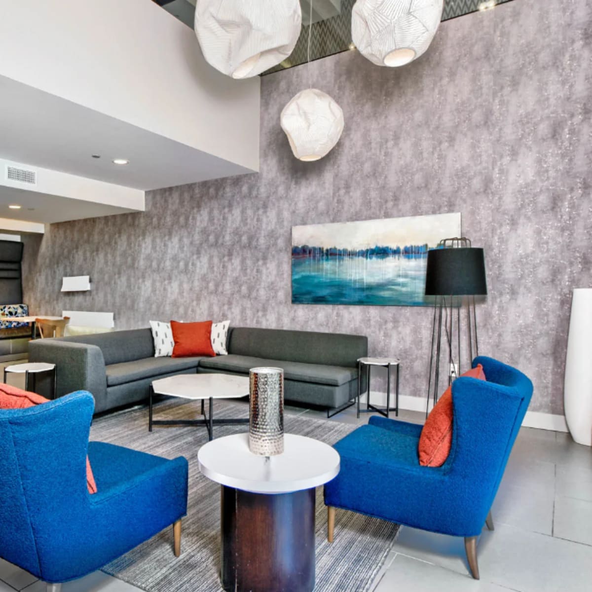 A modern lounge area with a gray wall, blue chairs, a sofa, a coffee table, and decorative light fixtures.
