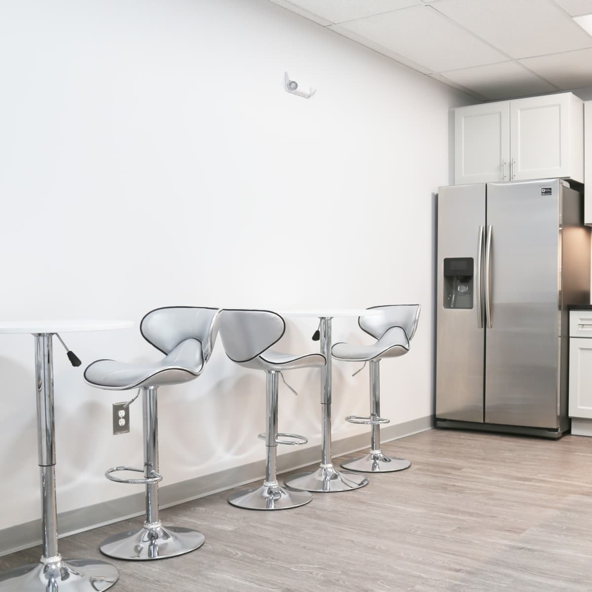 A modern kitchen area with a stainless steel refrigerator, a microwave, and four high-backed chrome stools arranged around two sleek white tables.