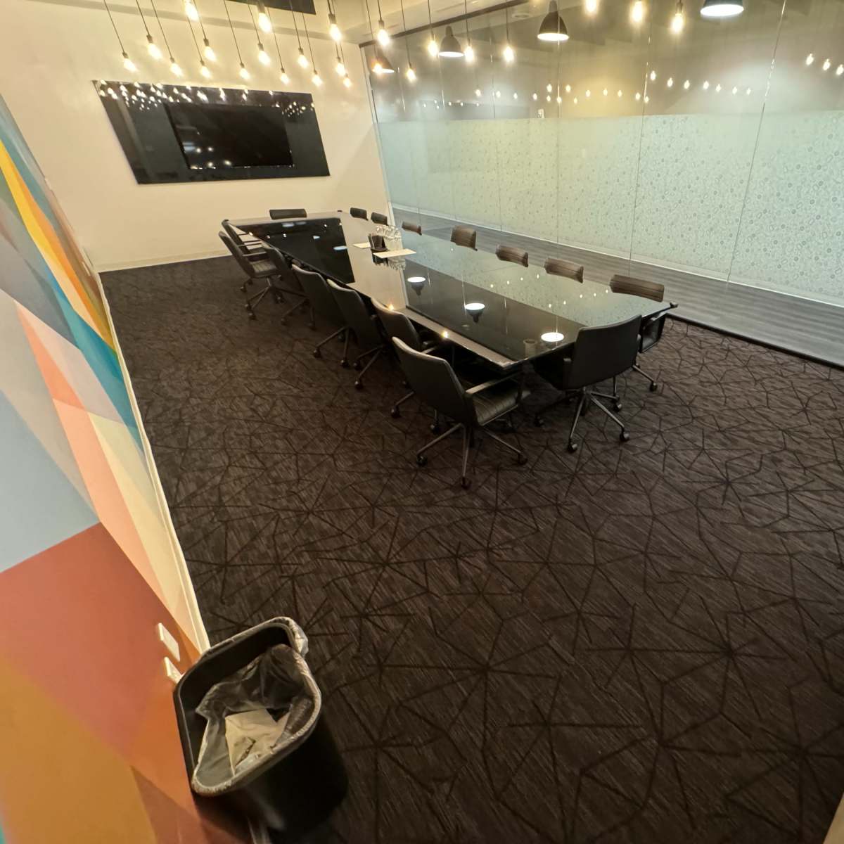 A large conference room features a long glass table surrounded by black chairs, with a television on one wall and decorative lighting overhead.