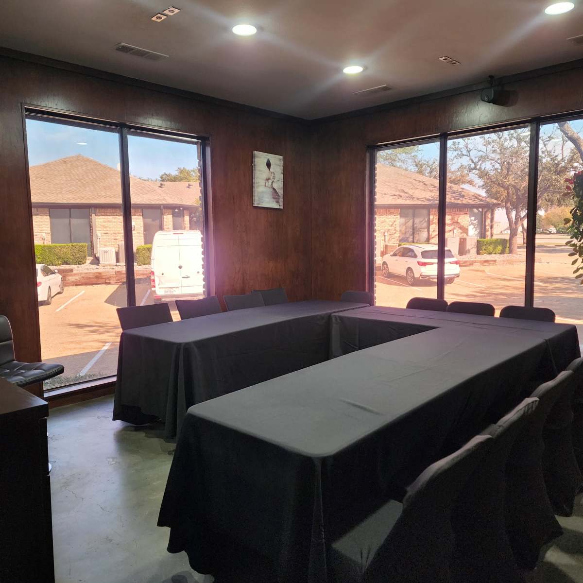A meeting room with a large rectangular table surrounded by chairs, illuminated by natural light coming through large windows.