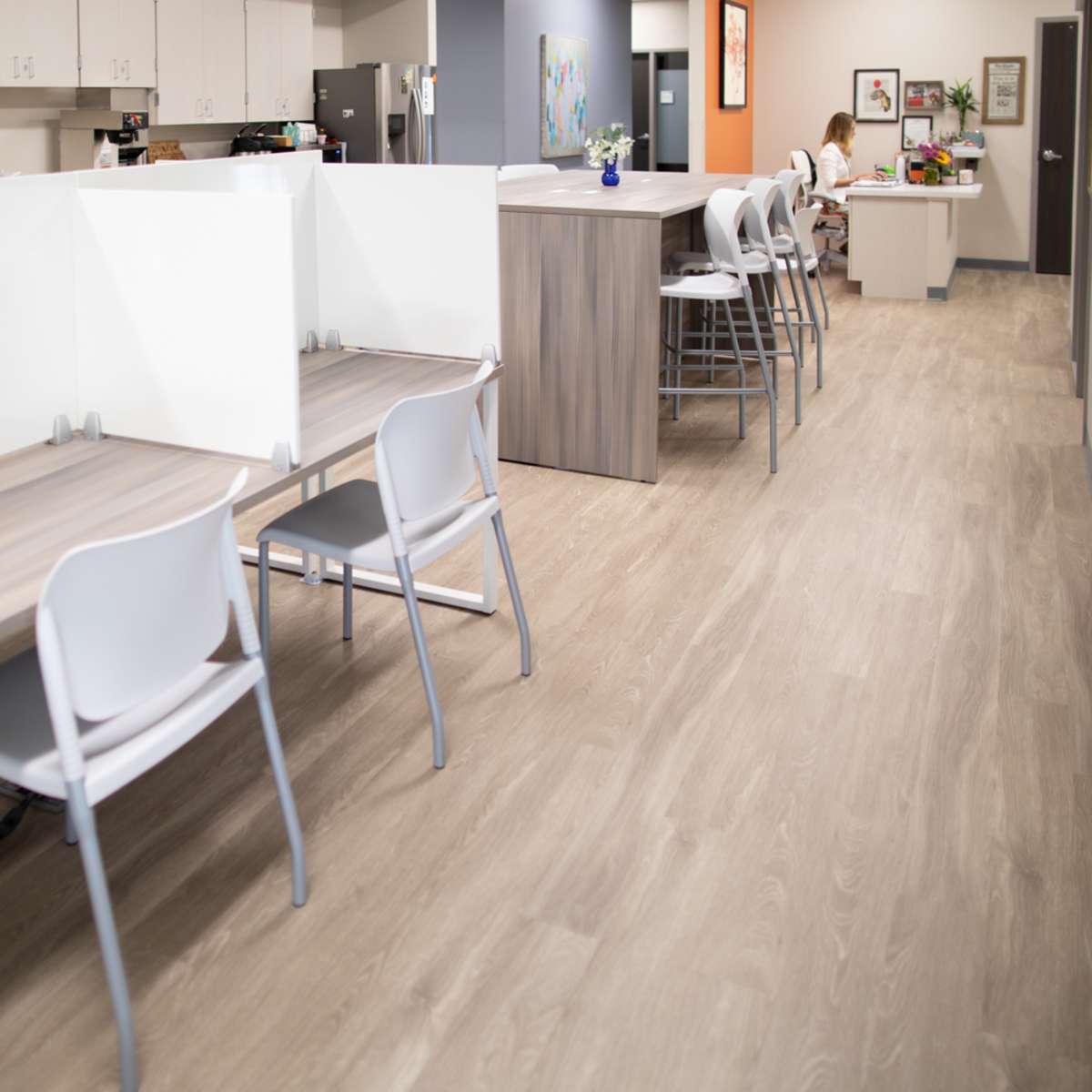 A modern office space with workstations separated by clear partitions, and a kitchenette area visible in the background.