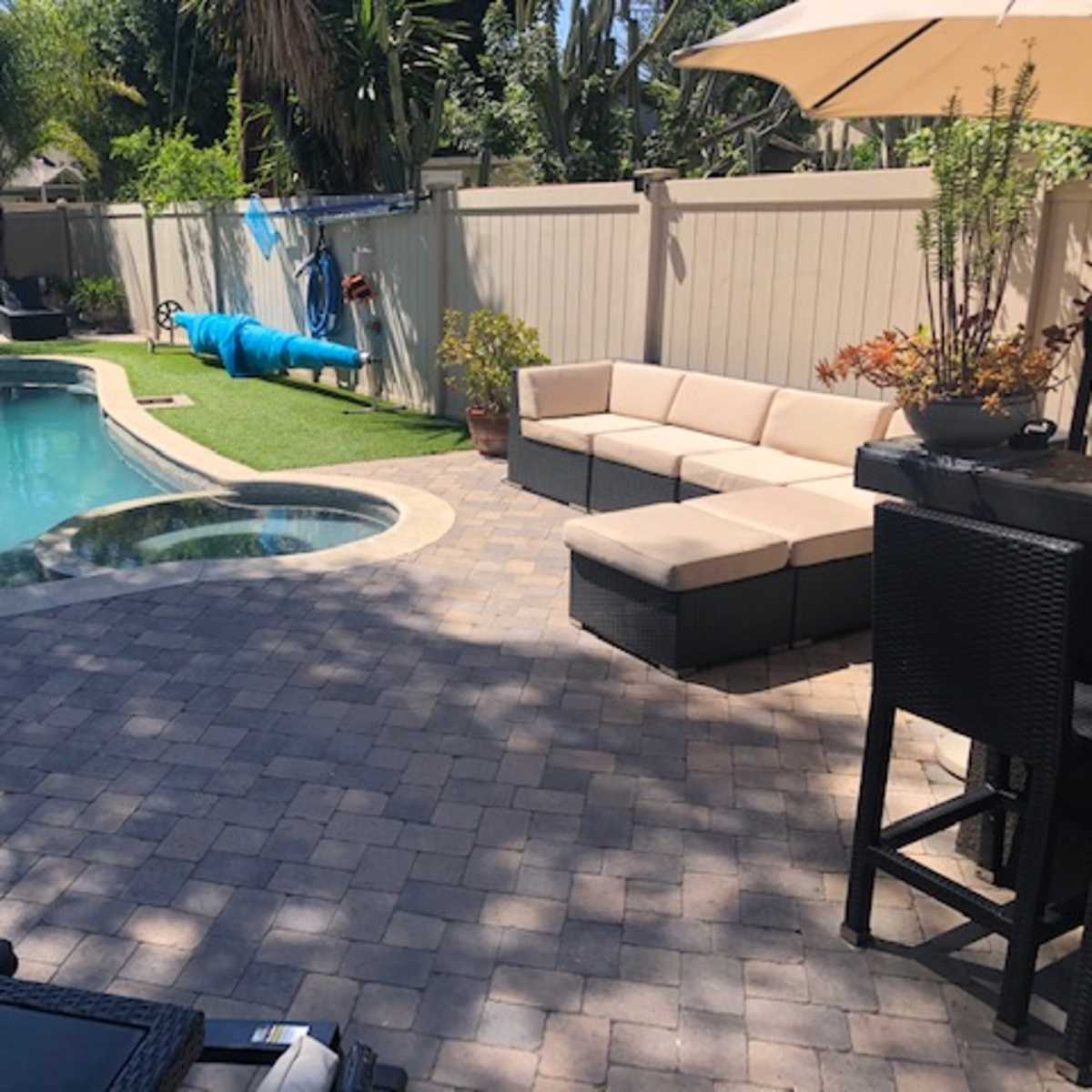 A landscaped backyard with a swimming pool, a spa, and a seating area featuring a sectional couch and tall bar stools under a patio umbrella.
