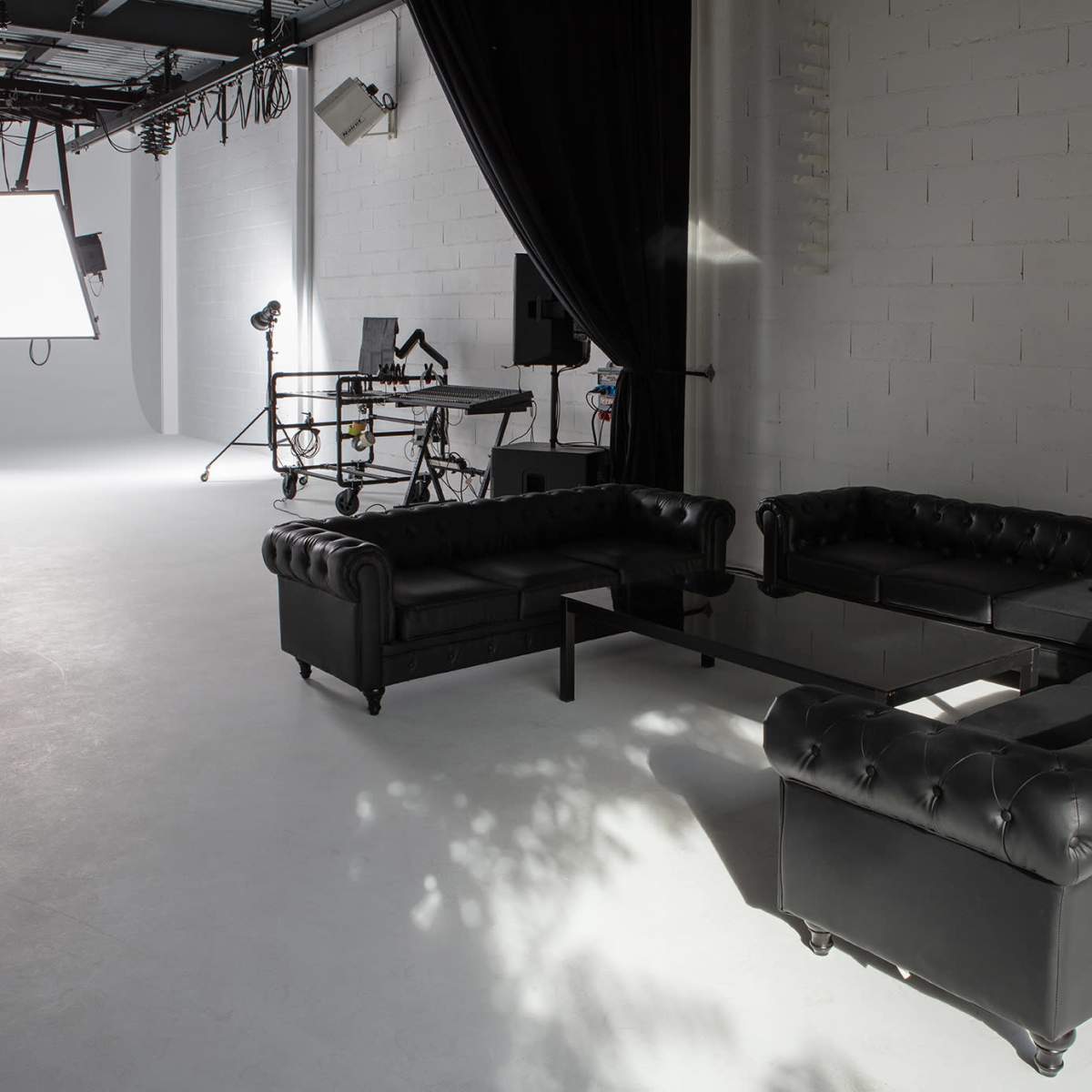 A modern studio setup featuring a black leather sectional couch, a glass coffee table, and various lighting equipment in a minimalist space.