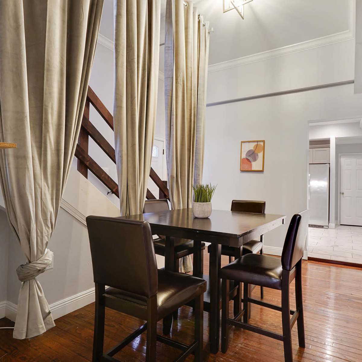 A dining area features a wooden table with four chairs, flanked by curtains and surrounded by a staircase and an open doorway leading to a kitchen.