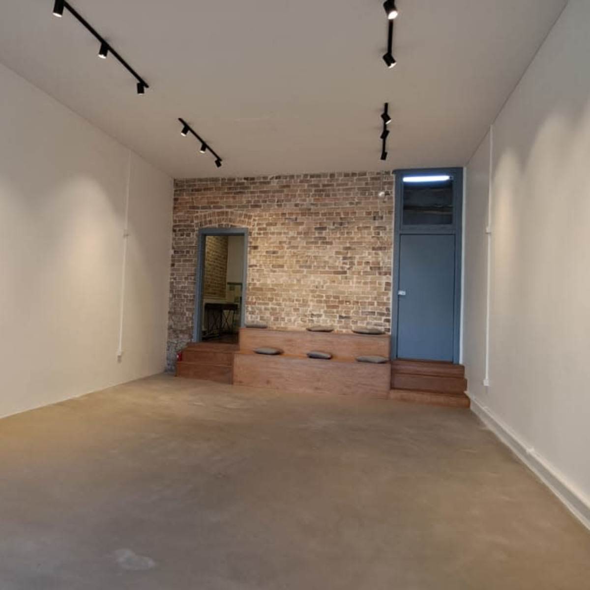 A spacious room with a minimalistic design featuring a desk with a chair, exposed brick wall, and steps leading to a smaller area at the back.