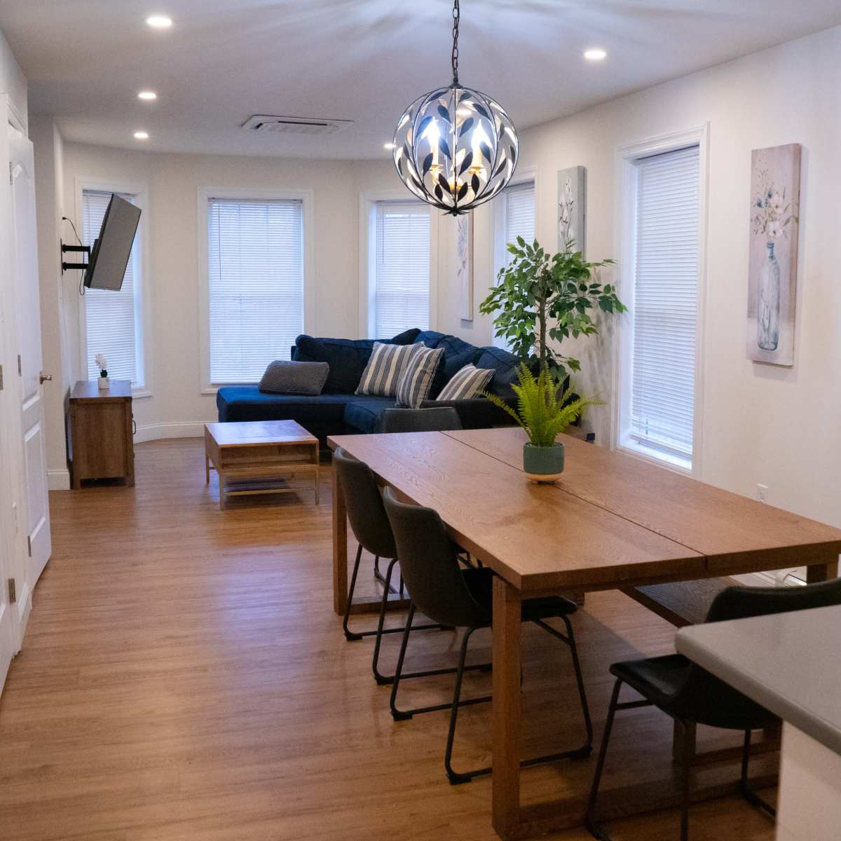 A modern living space features a dining table, chairs, a sofa, and large windows with natural light.