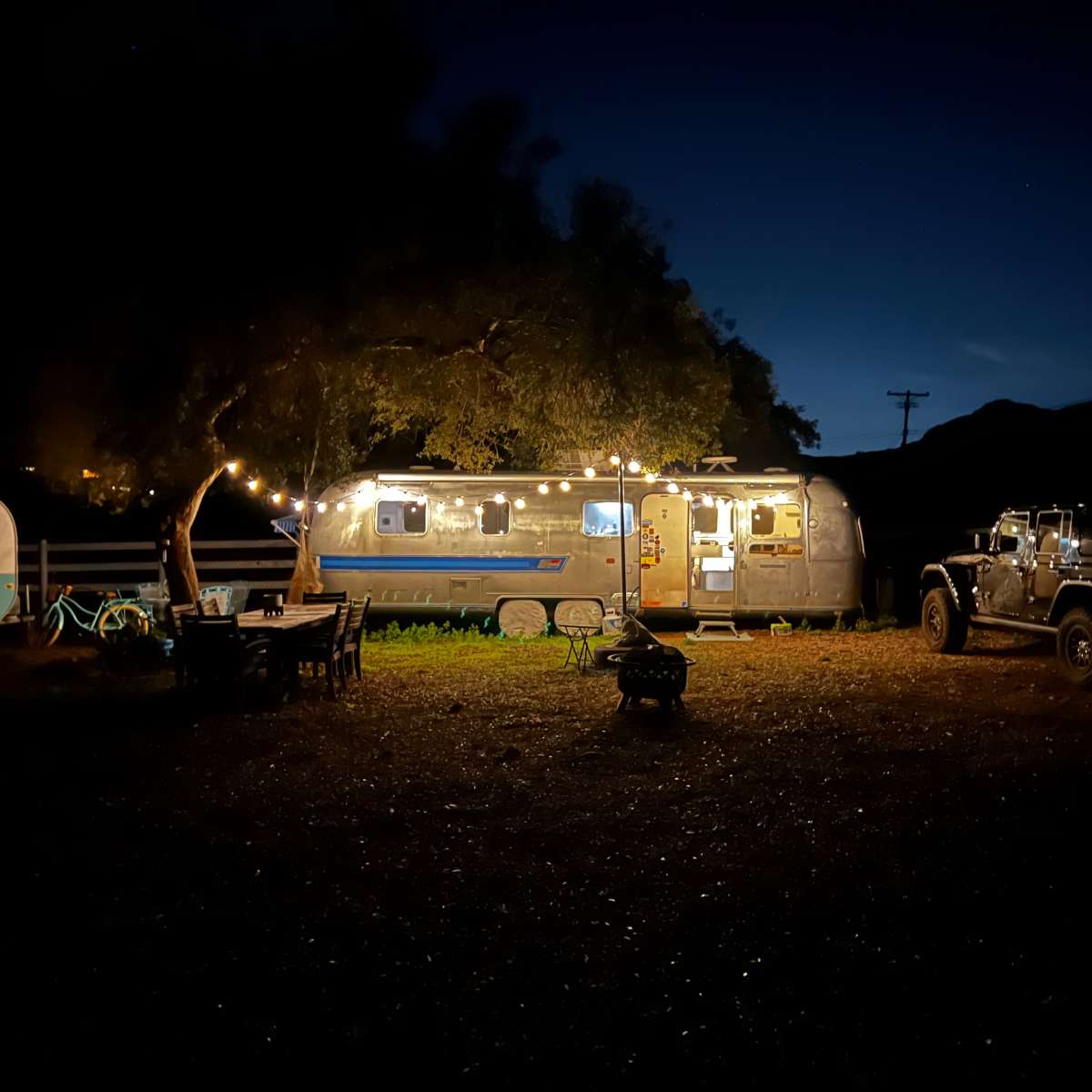 An Airstream trailer is illuminated at night with string lights, surrounded by outdoor seating and a Jeep nearby.