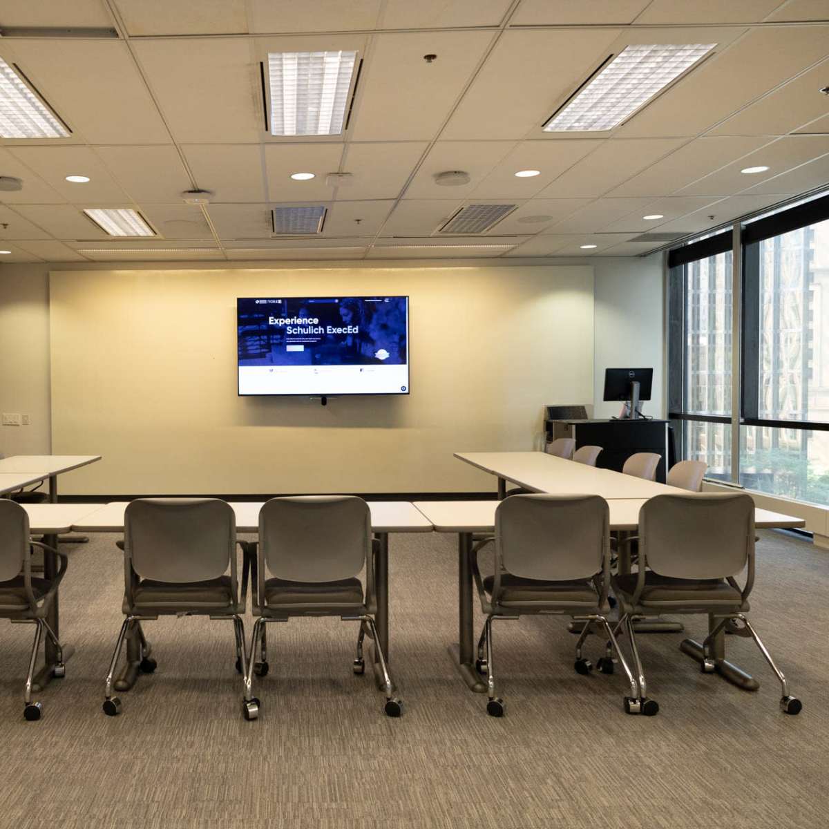A modern conference room features a central table surrounded by chairs, with a large screen mounted on the wall.