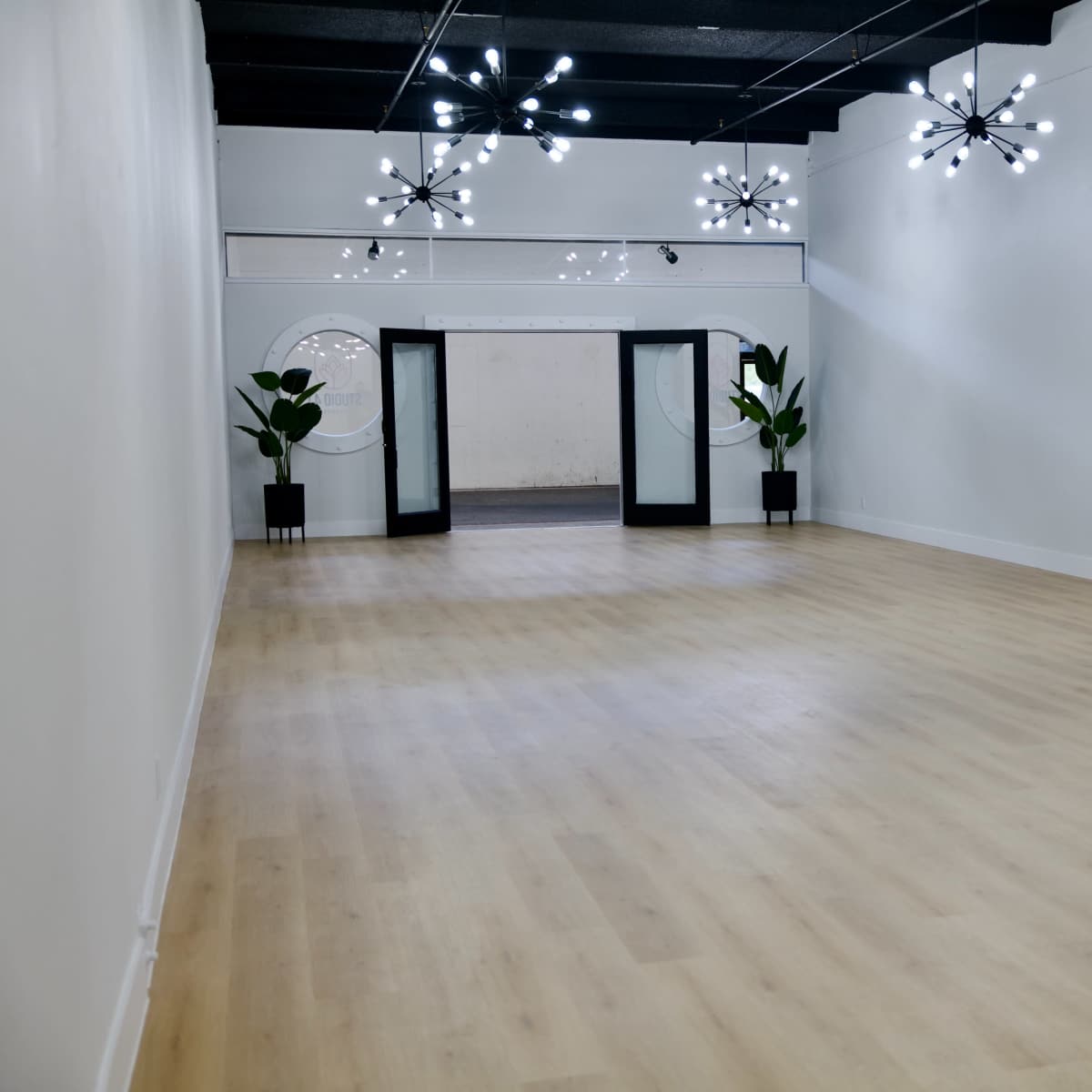A spacious, empty room with light wooden flooring, featuring modern chandeliers and two large glass doors leading to another area.