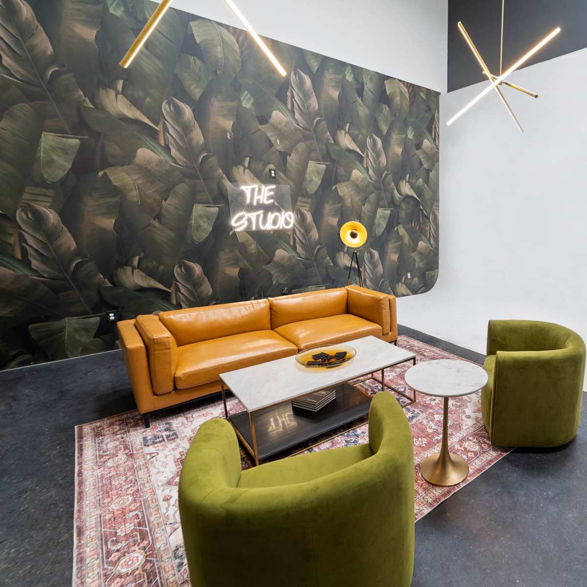 A modern lounge area features a yellow leather sofa, two green velvet chairs, a marble coffee table, and a wall decorated with a leafy mural.