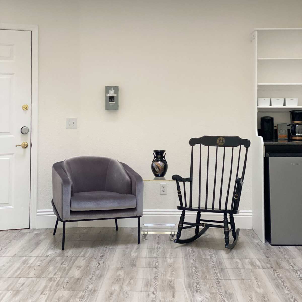 A gray armchair and a black rocking chair are placed against a light-colored wall in a room with a small kitchenette visible nearby.