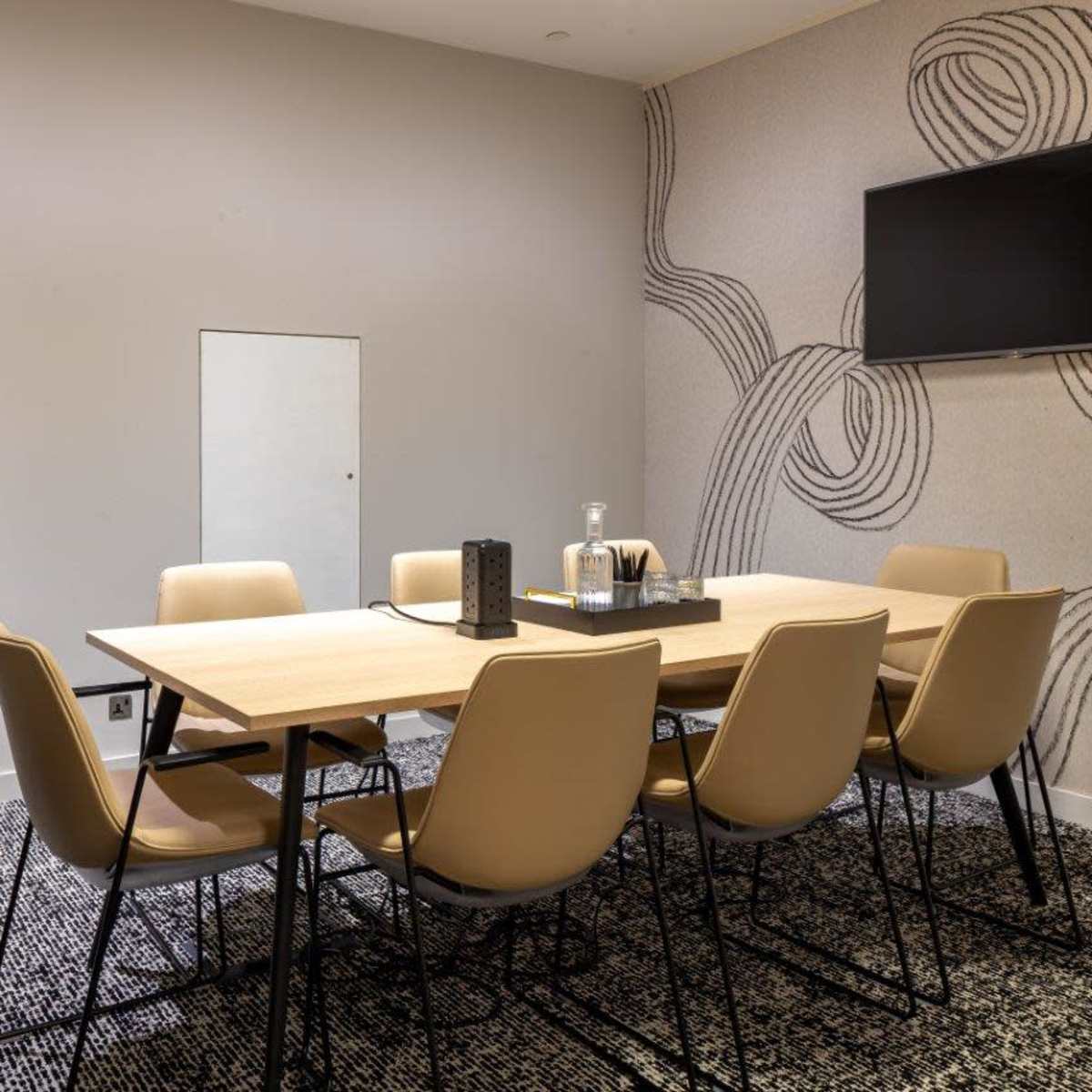A modern conference room features a large wooden table surrounded by six beige chairs, with a wall-mounted television and decorative wallpaper.