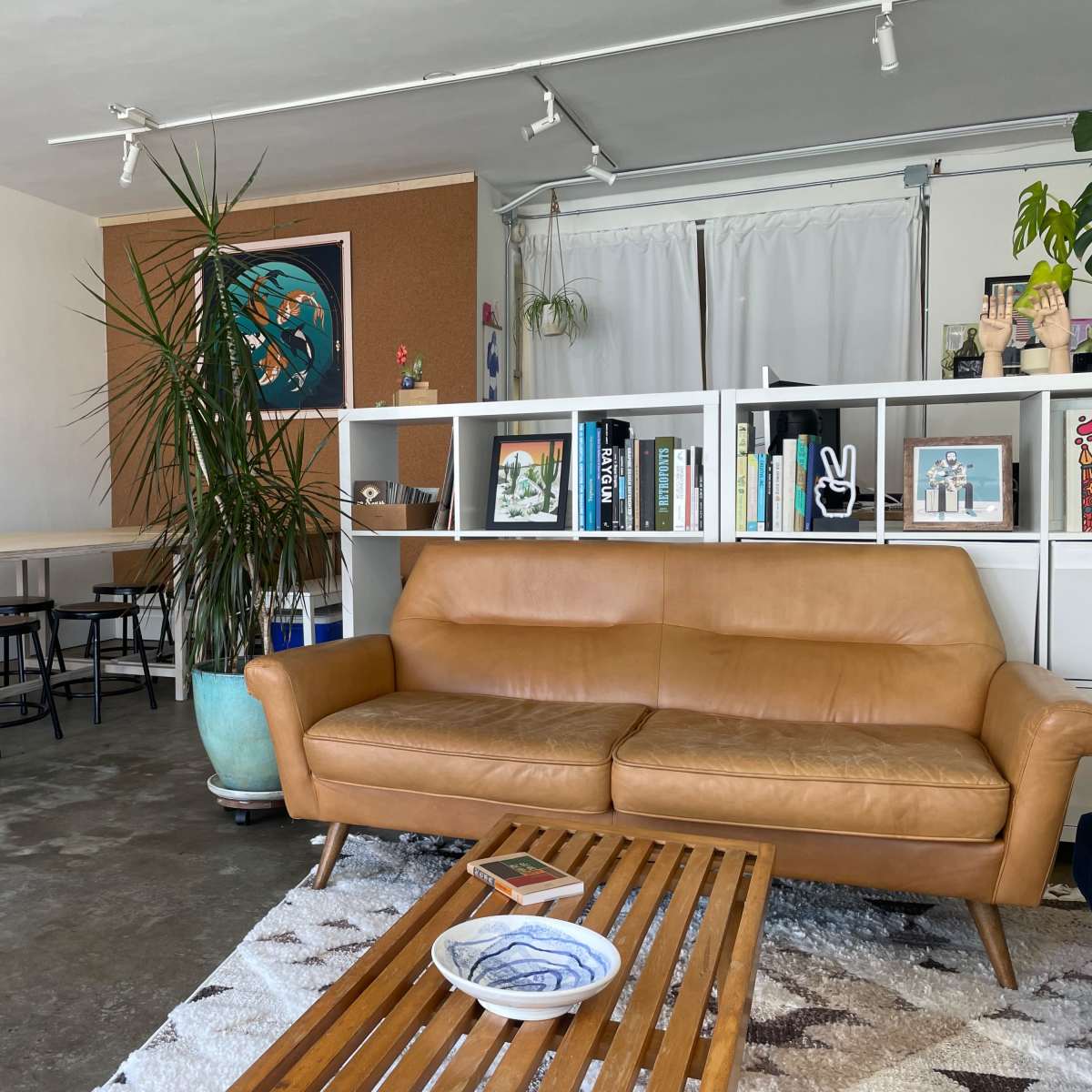 A modern living space with a brown leather sofa, a wooden coffee table, and a white bookshelf adorned with books and decor, set against a backdrop of wall art and a plant.