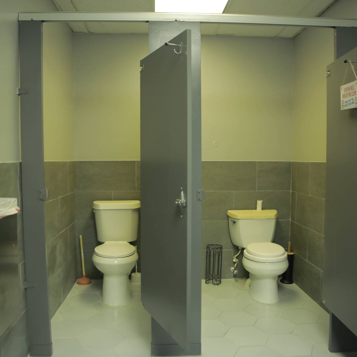 A public restroom with three toilet stalls, two of which are occupied, featuring gray partitions and tiled floors.