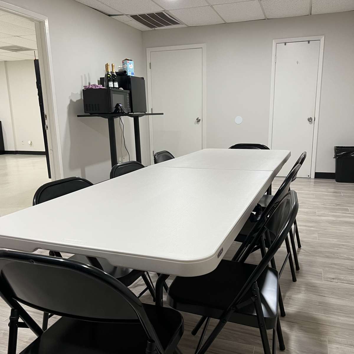 A rectangular table surrounded by black folding chairs is set up in a well-lit room with a door and a small shelf against the wall.