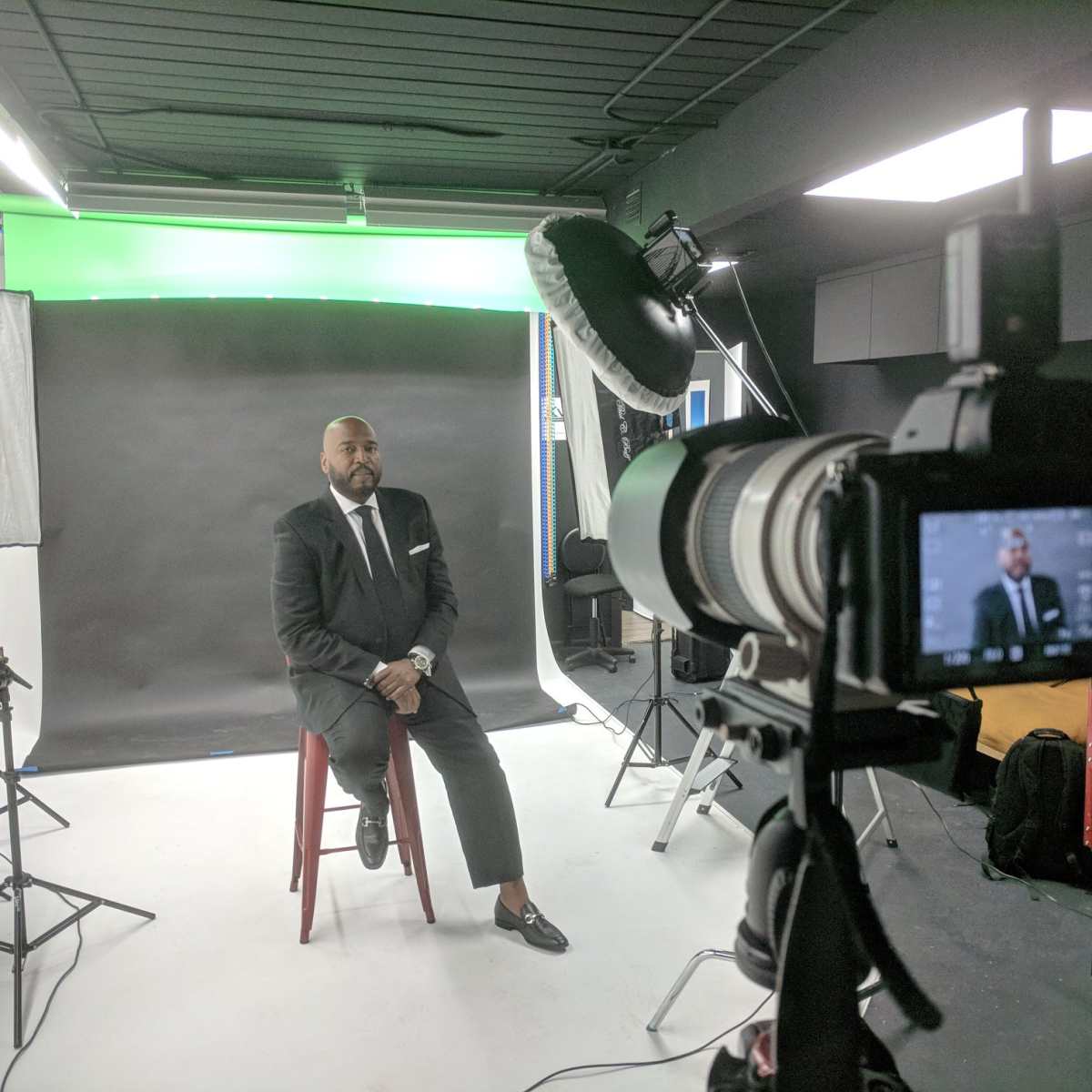 A man in a suit sits on a stool in front of a professional photography setup with multiple lights and a green backdrop.