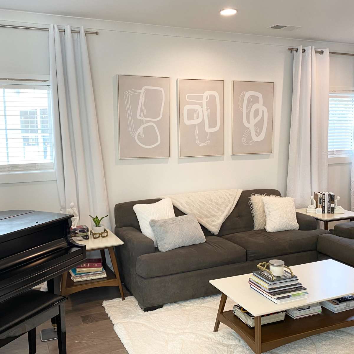 A modern living room featuring a dark sectional sofa, a coffee table with books and a plant, a grand piano in the corner, and three framed abstract artworks on the wall.