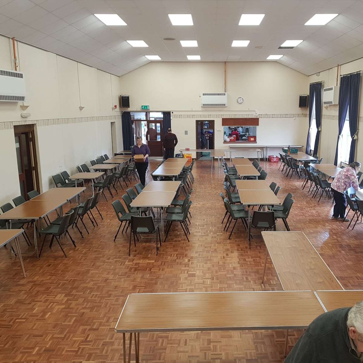 A spacious community hall with rows of tables and chairs set up for an event, featuring wooden flooring and large windows with curtains.