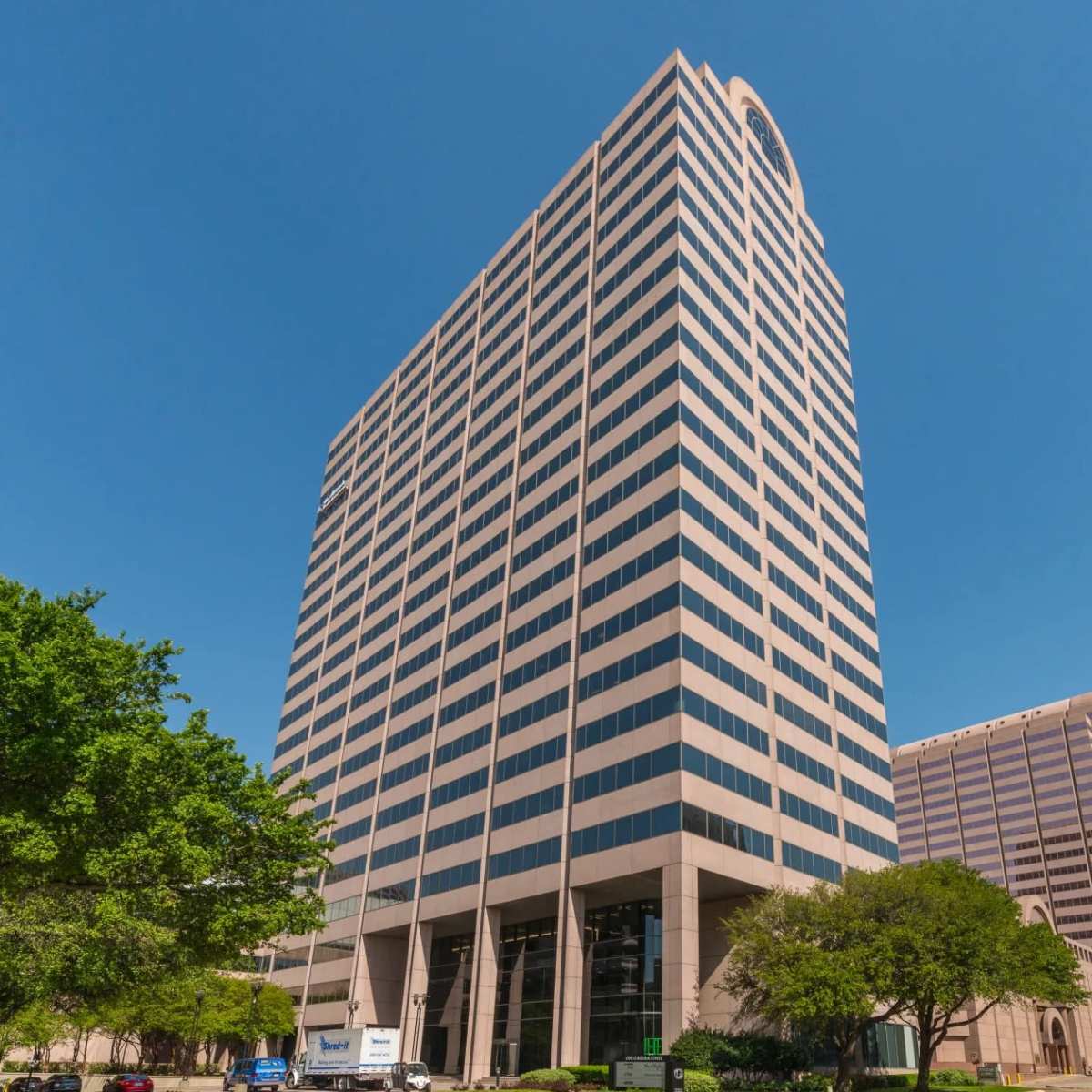 A tall office building with a modern design stands against a clear blue sky, flanked by trees and adjacent structures.