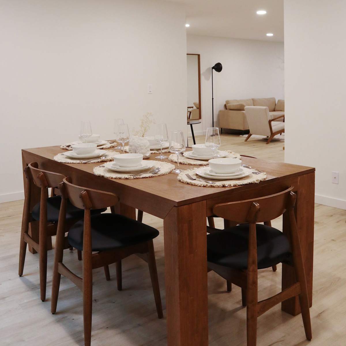 A wooden dining table is set with dishes and glasses, surrounded by chairs, while a living area is visible in the background.