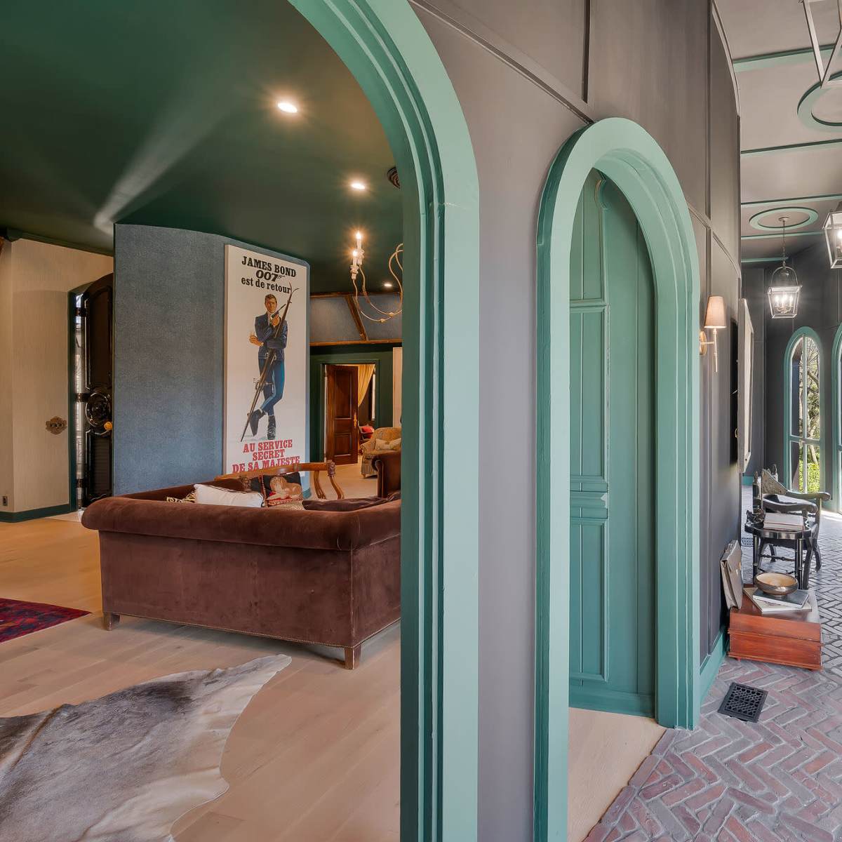 A hallway with curved arches, featuring dark walls and green trim, leading to a living area with a large poster and a brown sofa.