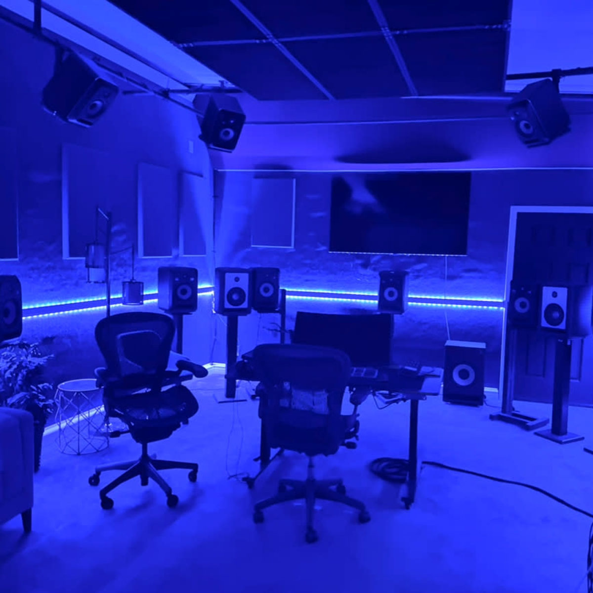 A music studio features multiple speakers, a central desk with chairs, and a large screen illuminated by blue lighting.