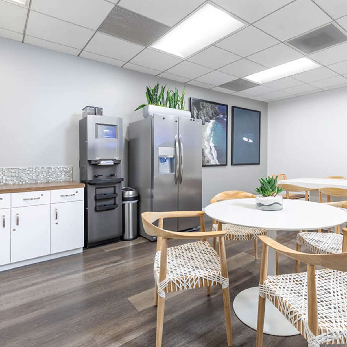 A modern break room with a round white table surrounded by wooden chairs, a refrigerator, and a kitchenette with storage cabinets.