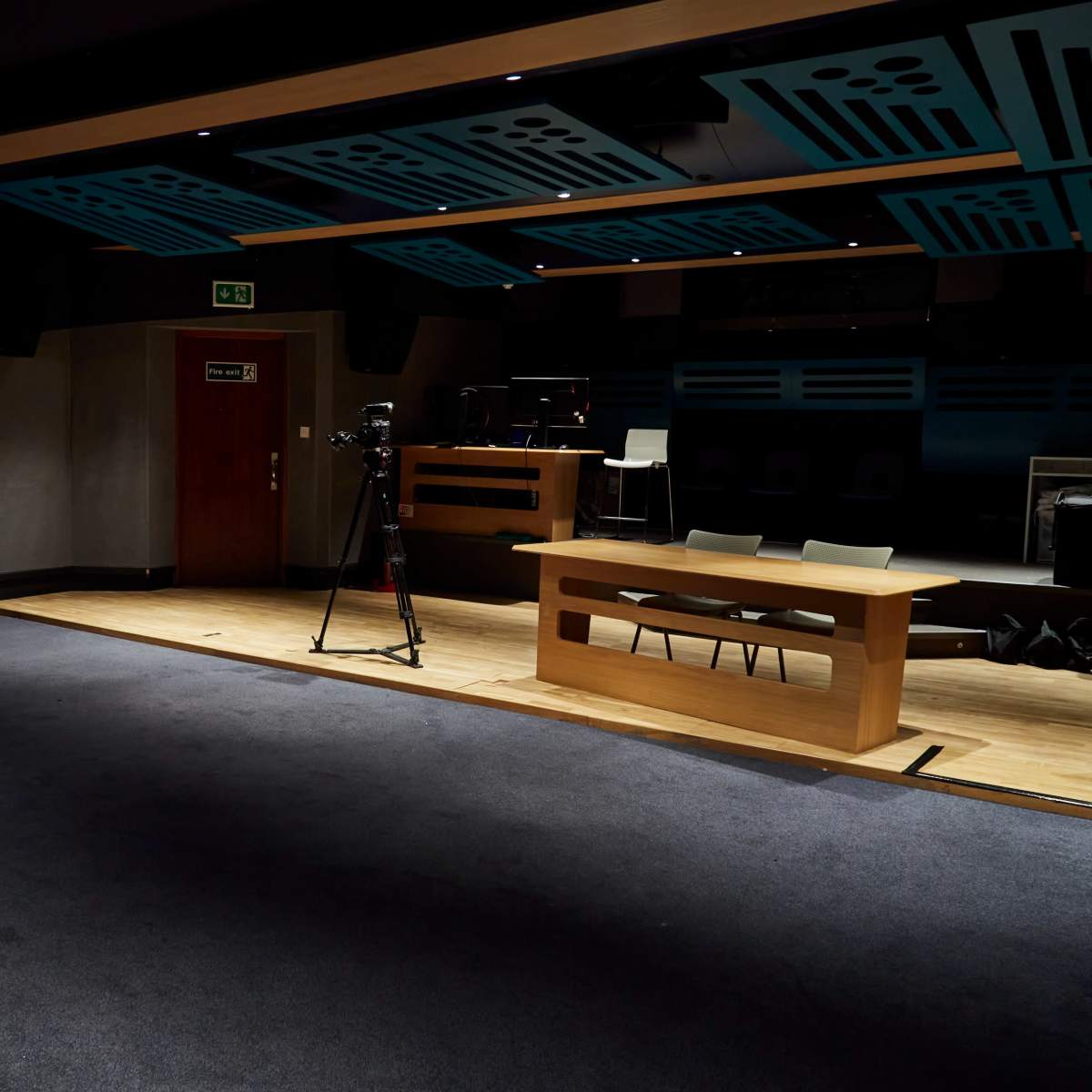 A dimly lit studio space featuring a single white chair, a wooden table, and a camera on a tripod.