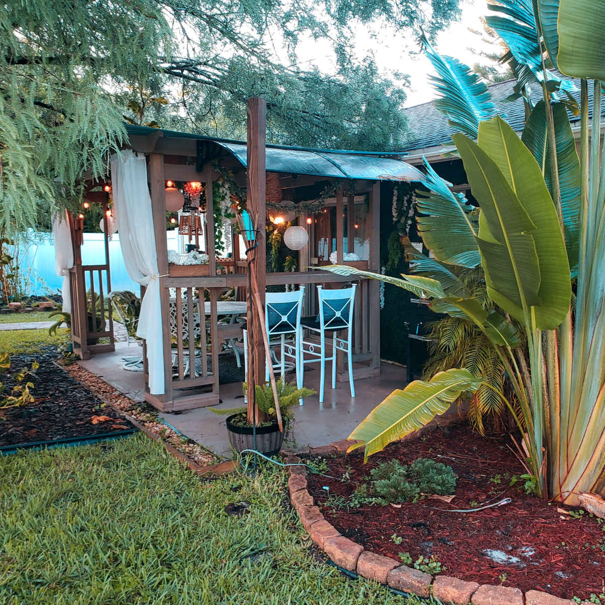 A wooden gazebo with a table and chairs is surrounded by lush greenery and tropical plants in a residential yard.