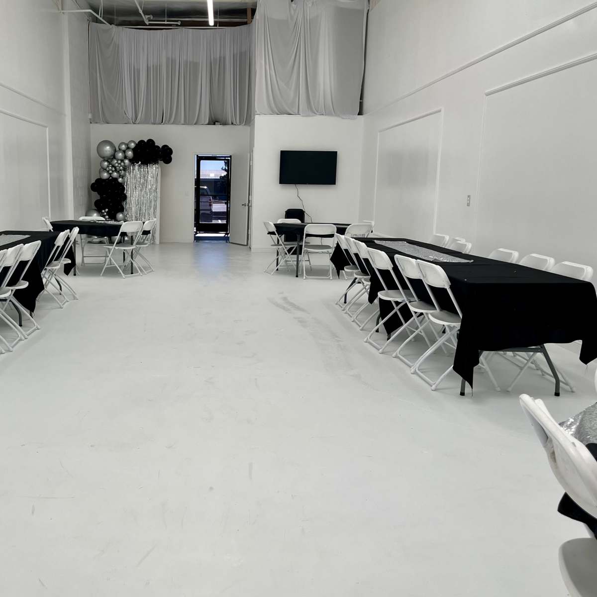 A spacious, empty room set up with black tablecloths over white folding tables arranged in two rows, with a black and white decorative arrangement in the corner.
