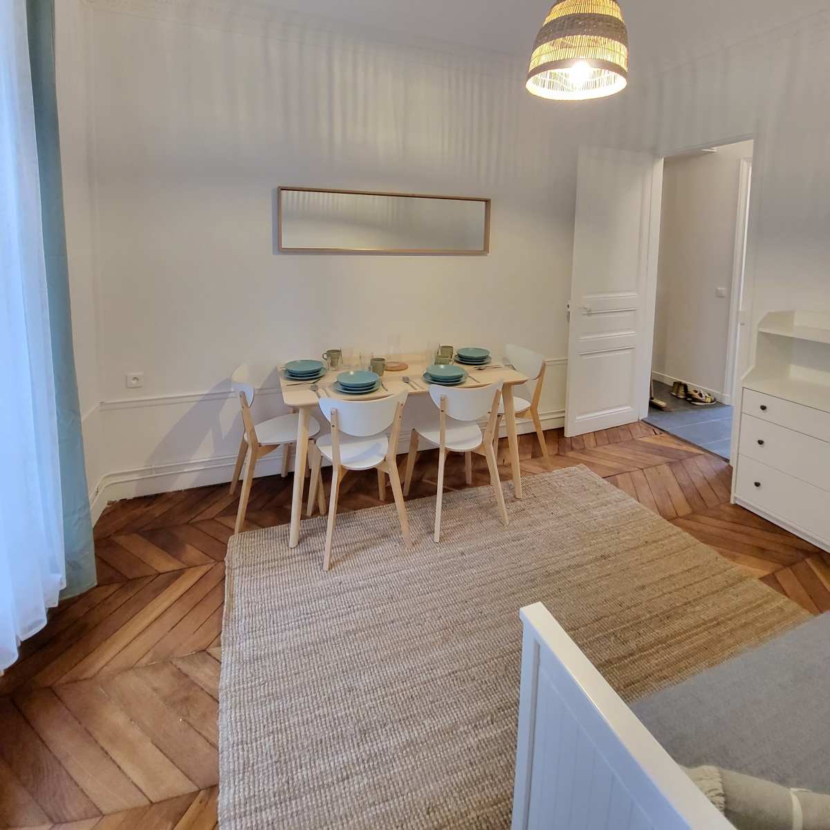 A dining area with a light-colored table and four chairs is set against a wall with a framed picture, illuminated by a pendant light.