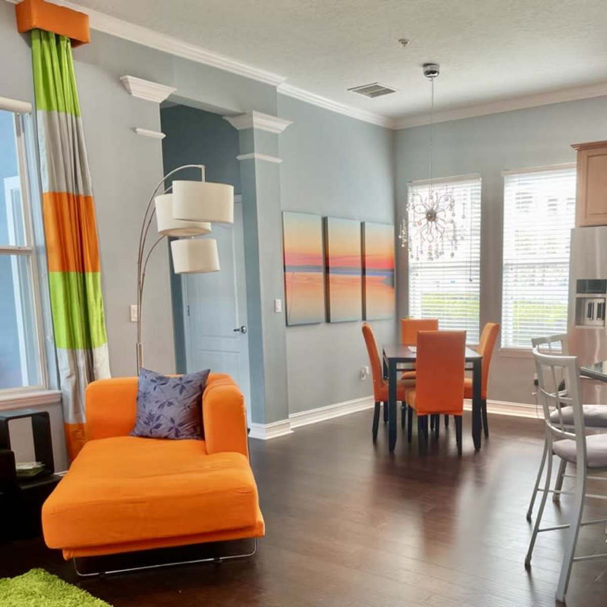 A modern living space with an orange sofa, a dining area with orange and gray chairs, and a kitchen with wooden cabinets and stainless steel appliances.
