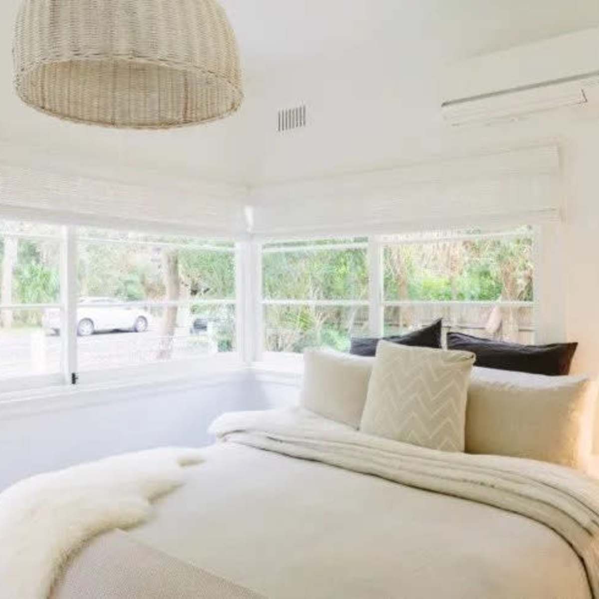 A bright bedroom with a bed covered in neutral bedding, large windows letting in natural light, and a woven pendant lamp hanging overhead.