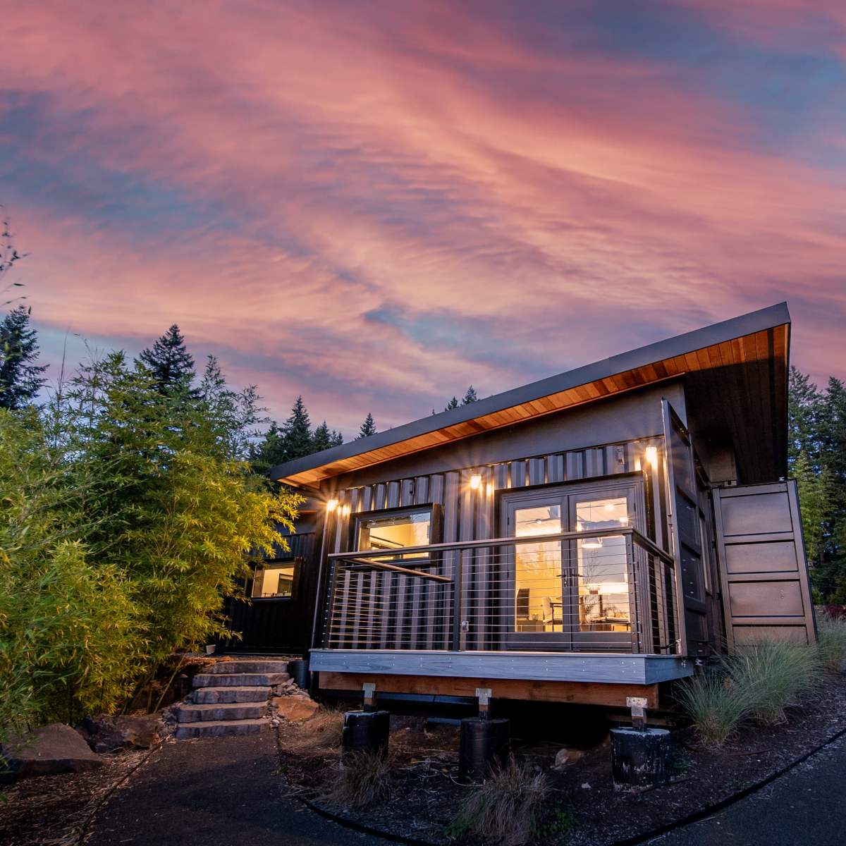 A modern cabin with large windows and a deck is illuminated under a colorful sunset sky, surrounded by trees and landscaped greenery.