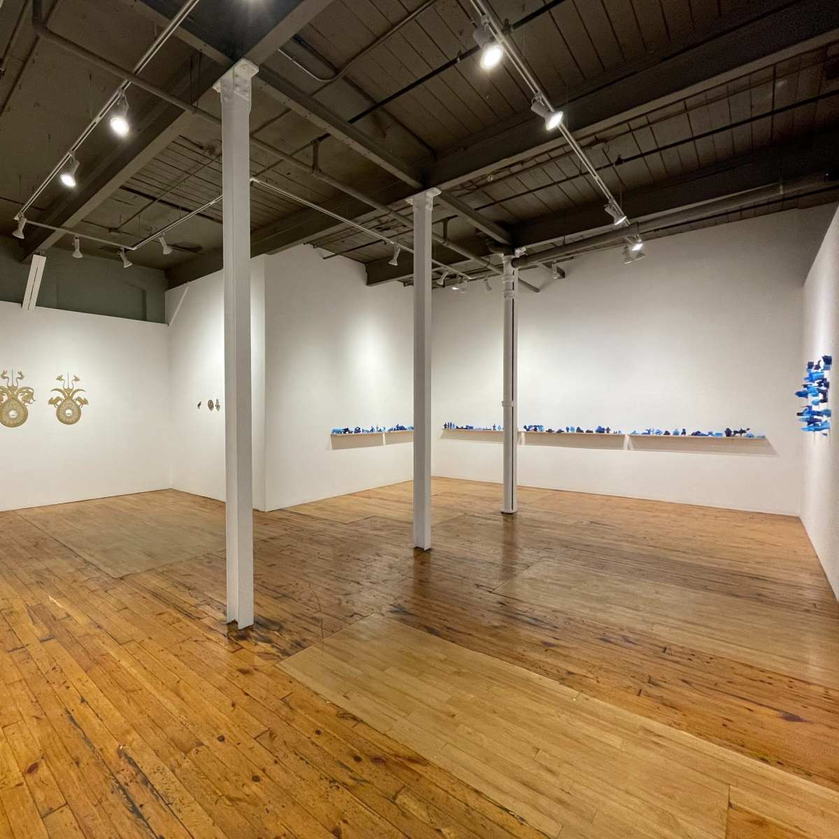 A spacious gallery with wooden flooring and white walls displaying various artworks, including two gold circular pieces on the left and a line of blue items on the right.
