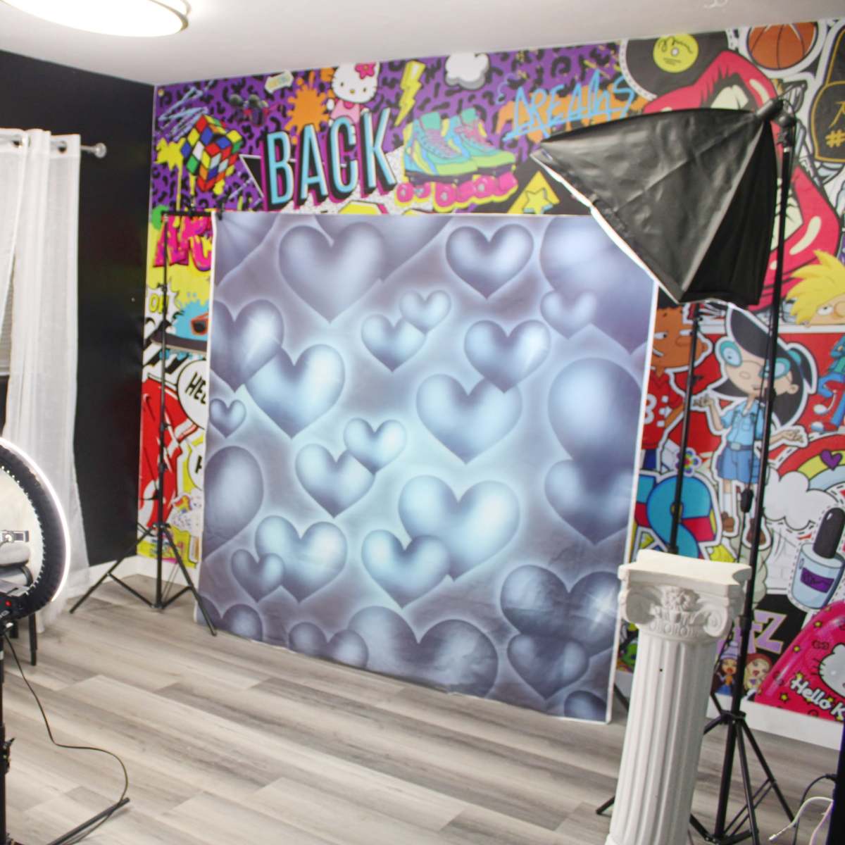A brightly decorated photography studio features a heart-patterned backdrop, a ring light, and a large window with sheer curtains.