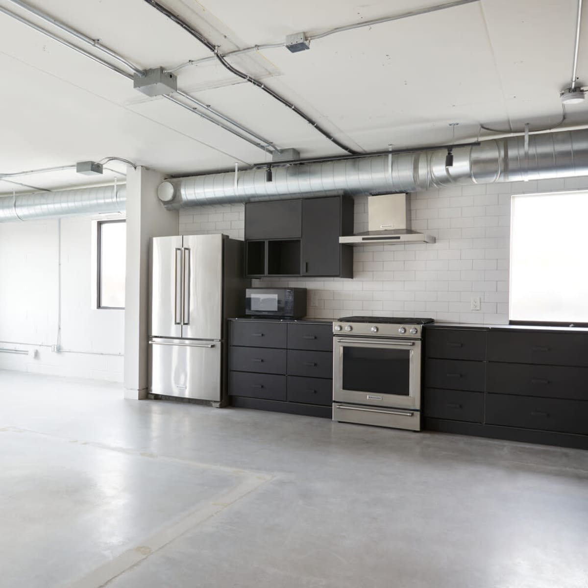 A spacious, modern kitchen area with stainless steel appliances and concrete flooring, featuring large windows on two walls.