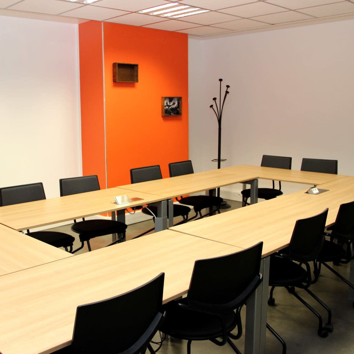 A conference room with a rectangular table arrangement surrounded by black chairs, featuring an orange accent wall and minimal decor.