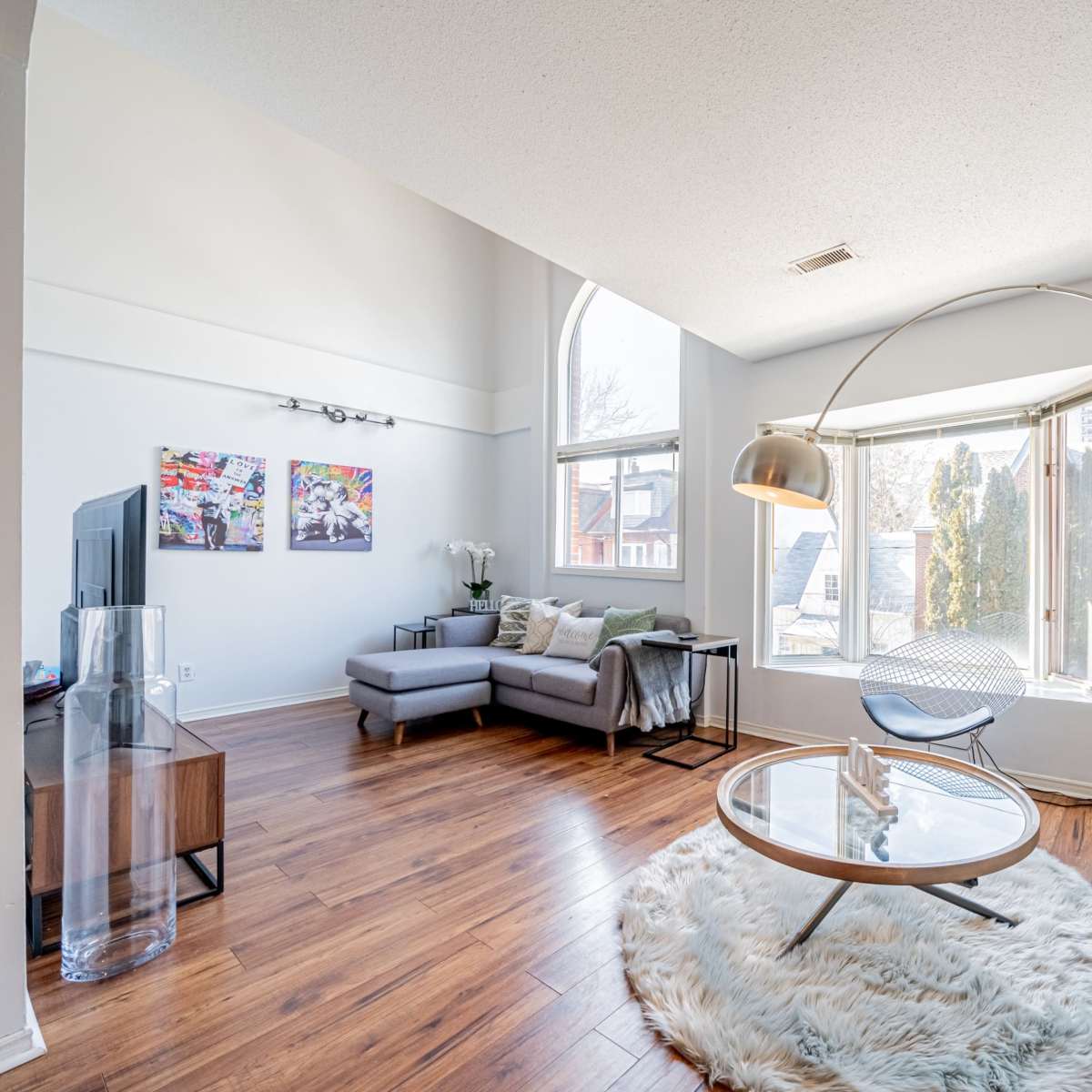 The living room features a gray sectional sofa, a round coffee table, an arc floor lamp, and large windows allowing plenty of natural light.