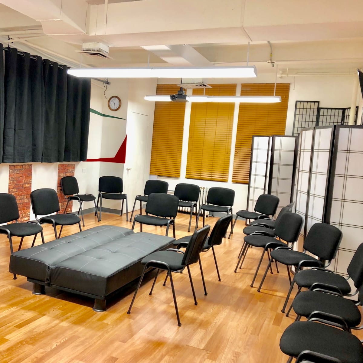 A room arranged with a circular seating layout of black chairs around a dark sofa on a wooden floor, with black curtains and a folding screen in the background.