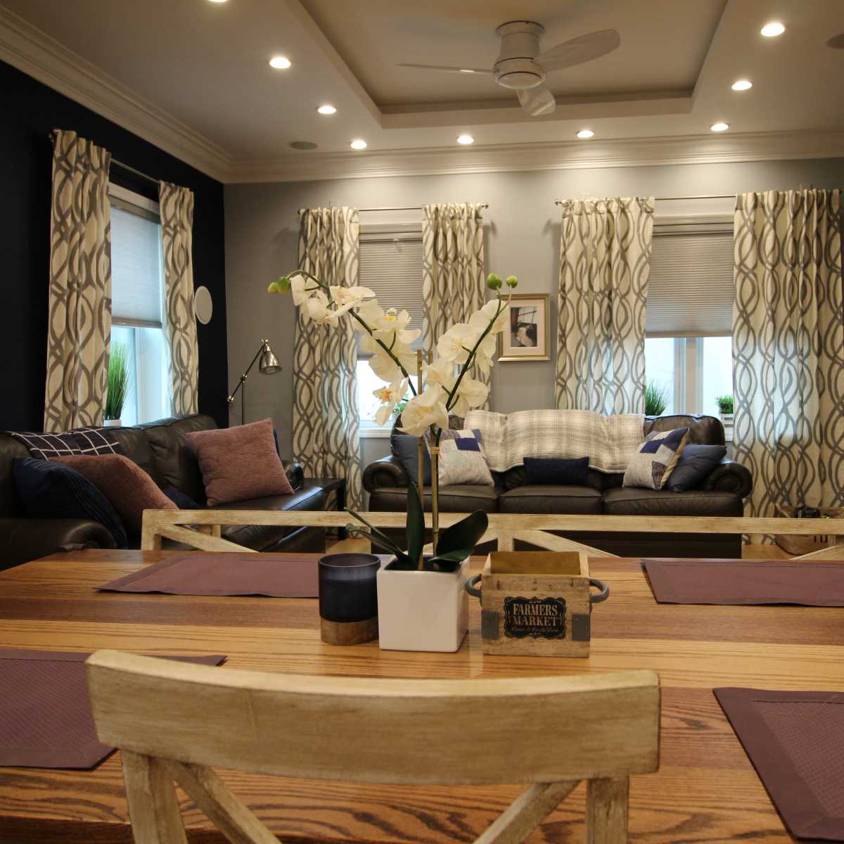 A dining area with a wooden table and chairs is set in front of a living room featuring dark walls, light curtains, and two sofas.
