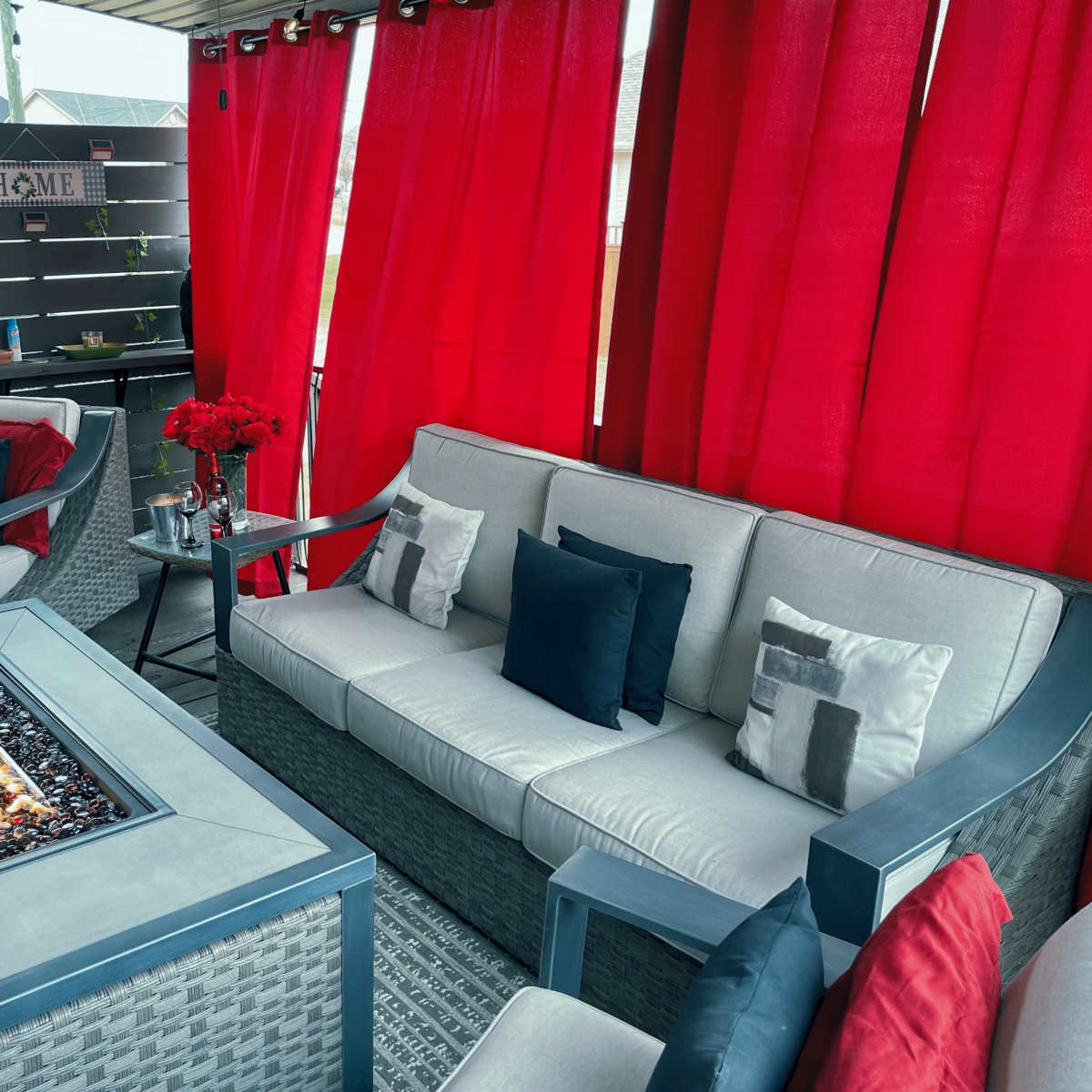 A cozy outdoor seating area features a fire table with glass stones, surrounded by a sofa and decorative pillows, against a backdrop of red curtains and a wooden wall.