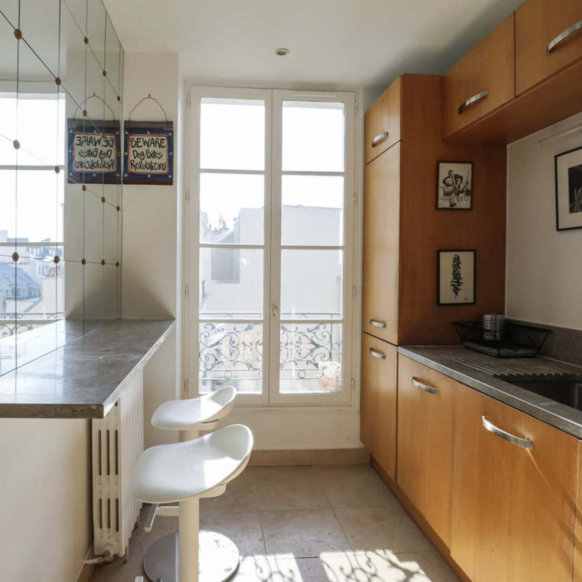 A modern kitchen features wooden cabinetry, a stainless steel sink, and two white stools at a counter near a window with a view.