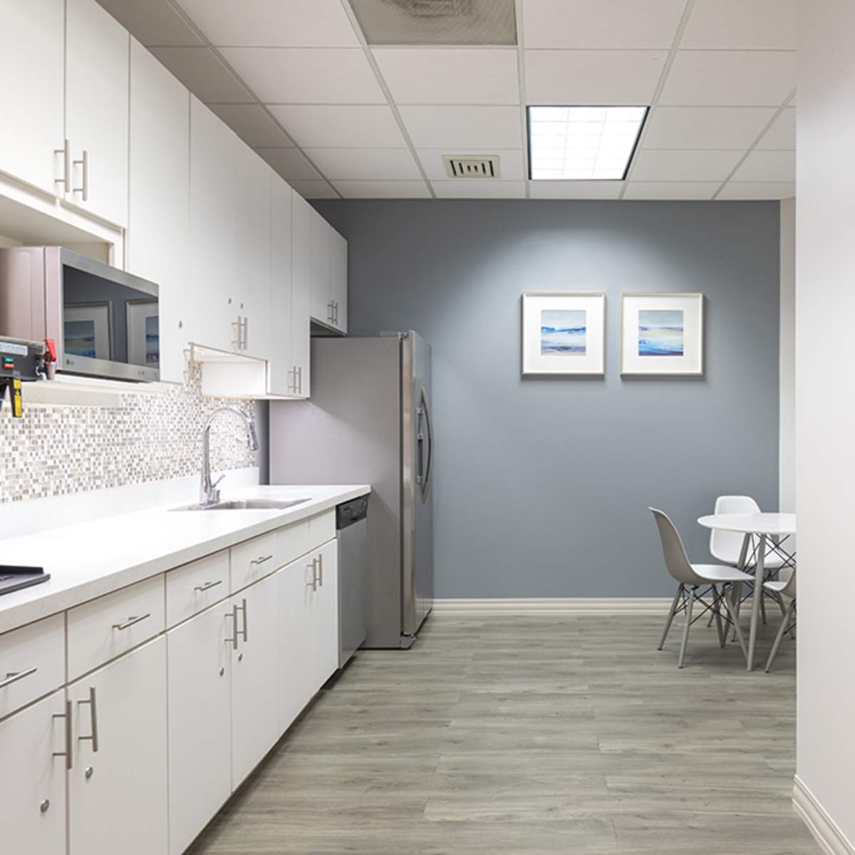 A modern kitchen area with white cabinetry, stainless steel appliances, a small dining table, and light grey walls adorned with two framed pictures.